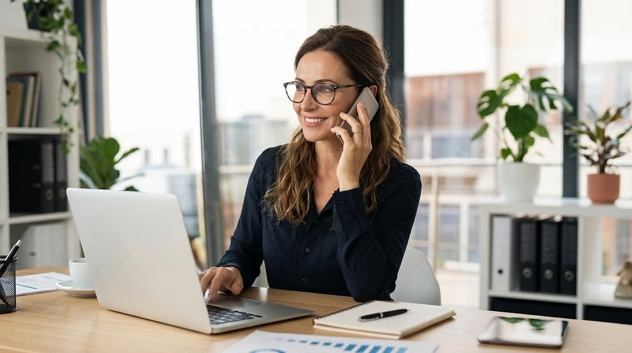 Una professionista sorride al telefono mentre lavora al portatile nel suo studio